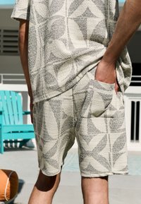 Lekki, fakturowany zestaw do noszenia w domu w kolorze beżowym z geometrycznym wzorem. Koszula ma krótkie rękawy; szorty posiadają boczne kieszenie.