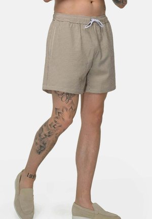 Man draagt beige-wit gestreepte shorts met een witte trekkoord, beige instapschoenen en zichtbare tatoeages op het linkerbeen.