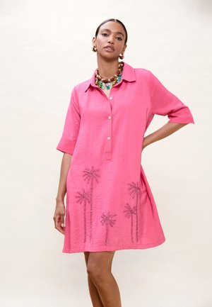 Mujer vistiendo un vestido rosa brillante con botones y bordado de palmeras en el dobladillo, complementado con pendientes gruesos dorados y un collar colorido.