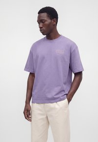 Homme portant un t-shirt lavande ample avec un petit texte blanc sur la poitrine et un pantalon beige clair, regardant sur le côté avec une main dans la poche.