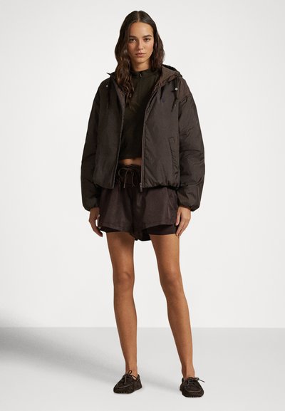 Γυναίκα που φοράει σκούρο καφέ μπουφάν με κουκούλα τύπου πάππου, αντίστοιχα σορτς, κοντό τοπ και σκουρόχρωμα παπούτσια μοκασίνι, στέκεται μπροστά σε απλό φόντο.