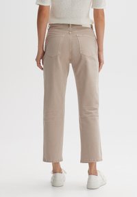 Mujer vestida con unos vaqueros rectos beige y zapatillas blancas, de pie frente a un fondo blanco liso, vista desde atrás.