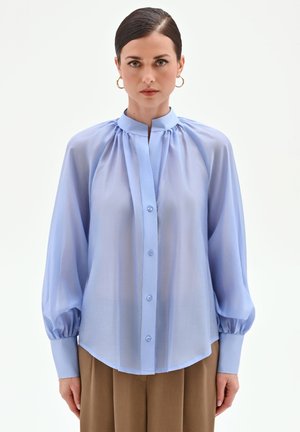 Donna con capelli scuri pettinati all'indietro che indossa orecchini a cerchio dorati, una camicetta azzurro chiaro trasparente con bottoni e maniche a sbuffo, e pantaloni marroni.