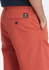 I pantaloni di cotone rossi presentano una texture liscia, tasche laterali e posteriori, un unico bottone nero sulla tasca posteriore e un'etichetta con il logo Timberland.
