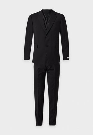 Un homme porte un costume noir deux pièces avec une chemise blanche et une cravate noire, sur un fond uni gris clair.