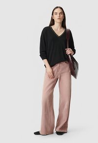 Schwarzer V-Ausschnitt-Pullover mit langen Ärmeln, kombiniert mit hellrosa Hosen mit weitem Bein. Das Modell hält eine dunkelviolette Tasche. Schwarze Schuhe mit flacher Sohle.