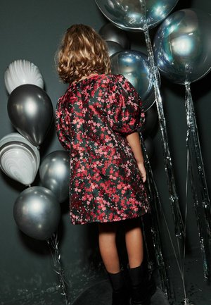 Robe noire à fleurs avec manches bouffantes, ornée de fleurs rouges et roses. Ballons argentés brillants et accents métalliques en arrière-plan.