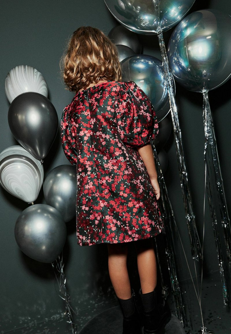 Robe noire à fleurs avec manches bouffantes, ornée de fleurs rouges et roses. Ballons argentés brillants et accents métalliques en arrière-plan.