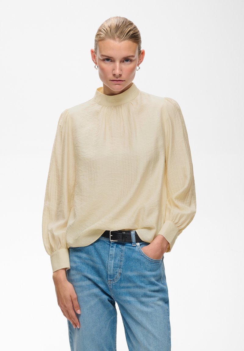 Femme aux cheveux blonds portant un chemisier crème à manches longues et un jean bleu, une main dans la poche, regardant devant avec une expression neutre.
