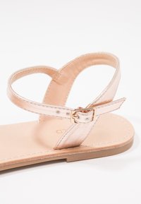 Vaaleanpunakultaiset litteät sandaalit, joissa on kapea nilkkaremmi, metallinen viimeistely, solkikiinnitys ja beige teksturoitu pohja. Minimalistinen muotoilu.