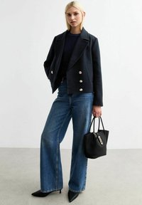 Giacca doppiopetto navy con bottoni argento, abbinata a jeans blu a gamba larga e una borsa tote nera. Tacchi neri a punta completano il look.