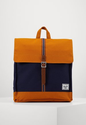 Sac à dos bicolore avec rabat et fond orange, corps bleu marine, sangle à boucle en cuir et poignée supérieure, posé sur une surface blanche.
