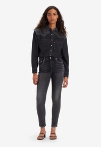 Camisa de mezclilla negra con botones a presión y dos bolsillos en el pecho, combinada con jeans ajustados negros de tiro alto con tachuelas plateadas. Sandalias negras.
