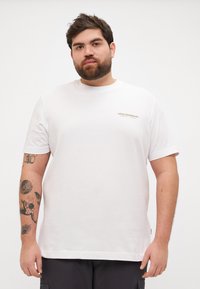 Camiseta de algodón blanca con mangas cortas, escote redondo y un pequeño logotipo dorado en el pecho. El corte es relajado y casual.