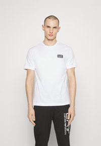 Camiseta de algodón blanca con mangas cortas, que presenta un pequeño parche de logo negro en el pecho. Textura suave, cuello redondo, diseño sin adornos.