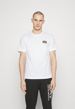 Camiseta de algodón blanca con mangas cortas, que presenta un pequeño parche de logo negro en el pecho. Textura suave, cuello redondo, diseño sin adornos.