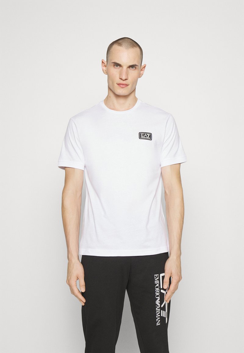 Camiseta de algodón blanca con mangas cortas, que presenta un pequeño parche de logo negro en el pecho. Textura suave, cuello redondo, diseño sin adornos.