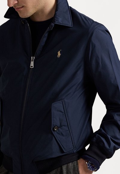 Άνδρας που φοράει ναυτικό μπουφάν με φερμουάρ και κουμπωτή τσέπη, με χρυσό κεντητό λογότυπο παίκτη πόλο στο στήθος και μπλε ρολόι χειρός.