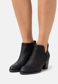 Bottes noires à la cheville en matériau tissé, avec une découpe arrondie, une fermeture éclair, et un talon carré. Texture lisse avec un léger éclat.