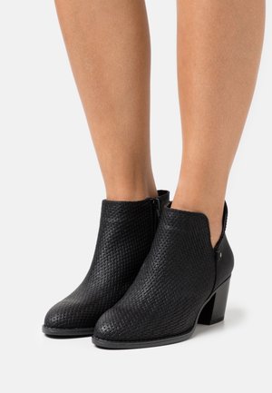Bottes noires à la cheville en matériau tissé, avec une découpe arrondie, une fermeture éclair, et un talon carré. Texture lisse avec un léger éclat.
