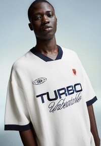 White short-sleeve sports jersey with navy accents, featuring “TURBO Unbeatable” text, ribbed texture, and a small logo on the chest.