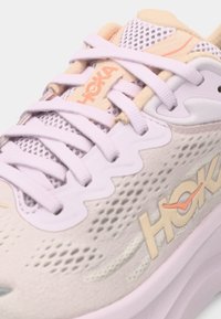 Μωβ και μπεζ αθλητικό παπούτσι με επάνω μέρος από δίχτυ, παχύ πάτο και λογότυπο HOKA, με ορατά κορδόνια και υφή στους λεπτομέρειες.