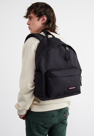 Sac à dos en tissu noir avec une poche zippée à l'avant, des bretelles ajustables et un patch logo. Porté par une personne portant un sweat-shirt de couleur claire.
