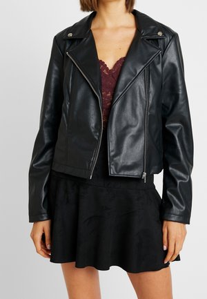 Kobieta w czarnej sztucznej skórzanej kurtce motocyklowej, nałożonej na ciemnoczerwoną koronkową bluzkę i czarną spódnicę, z rękami swobodnie opuszczonymi wzdłuż ciała.