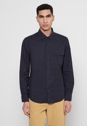 Navy long-sleeve shirt made of cotton, featuring a button-down collar, front pocket, and textured fabric, paired with beige pants.