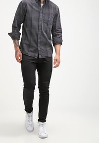 Mężczyzna idący, ubrany w ciemnoszarą koszulę na guziki, czarne spodnie, białe trampki oraz z tatuażami na lewym przedramieniu i dłoni.