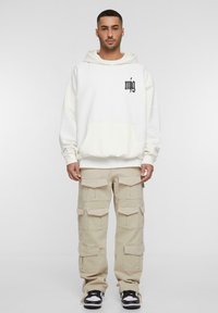 Sweatshirt à capuche blanc avec poche avant et logo noir, associé à un pantalon cargo beige ample avec de multiples poches, porté avec des baskets noires.