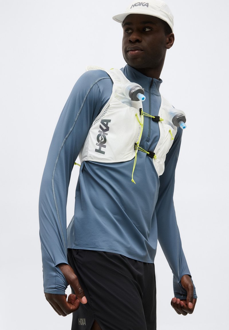 Hombre con camiseta deportiva azul de manga larga y pantalones cortos negros que lleva un chaleco de hidratación HOKA blanco con dos botellas de agua y gorra blanca.