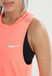 Camiseta sin mangas de Nike en un brillante color coral con una capa inferior negra. Presenta un cuello redondo y un detalle de malla transpirable en los hombros. Acento del logo en color plateado.