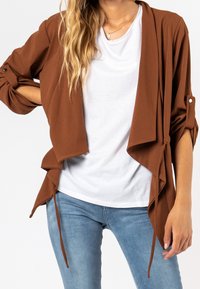 Brun lättviktsjacka med draperad design, öppen front och långärmar uppvikta, lagrad över en vit t-shirt och blå jeans.