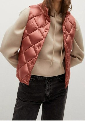 Kobieta w pikowanej kamizelce w kolorze przybrudzonego różu na beżowej bluzie z kapturem i czarnych dżinsach, z rękami za plecami.