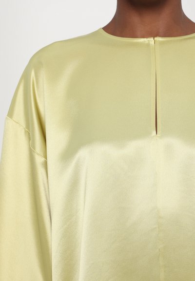 Top de color amarillo suave con un escote redondeado y una abertura en la parte posterior. Presenta hombros caídos y una textura lisa.