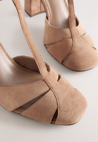 Brązowe zamszowe sandały na obcasie z krzyżującymi się paskami, zaokrąglonym noskiem i teksturowanym wykończeniem. Obcas typu słupek dla zapewnienia stabilności.