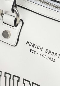 Bijela kožna torba s crnim detaljima, s patentnim zatvaračem i metalnim hardverom. Natpis "MUNICH SPORT BCN - EST.1939" otisnut sa strane.