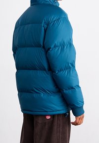 Turkos vadderad jacka med horisontella sömmar, hög krage och ribbade muddar. Märkbara detaljer inkluderar en dragsko vid fållen och en slät yta.