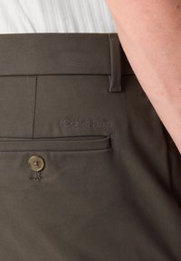 Donkerbruine Calvin Klein broek met een gladde textuur, voorzien van een knoopsluiting en een achterzak met geborduurd logo.