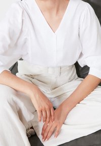 Hvid linnedbluse med V-udskæring og ballonærmer, parret med højtaljede hvide bukser med et bredt bælte. Hænderne hviler på skødet.