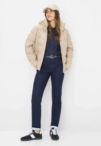 Vrouw in een beige pufferjas, marineblauw shirt, donkerblauwe spijkerbroek, witte en zwarte sneakers, en een beige baseballpet, staand tegen een witte achtergrond.
