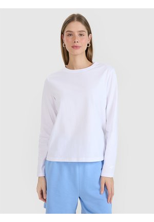 Joven mujer con una camisa blanca lisa de manga larga y pantalones azul claro, de pie frente a un fondo liso gris claro.