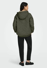 Olivzaļš kapucēts dūnu jaciņa ar gludu tekstūru un smalku rakstu, kombinēts ar melnām biksēm un melniem loaferiem.