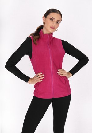 Fuchsiales Fleece-Westen mit hohem Kragen, durchgehendem Reißverschluss und Seitentaschen, getragen über einem schwarzen Langarmshirt und schwarzen Leggings.