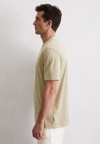Helles beige T-Shirt mit kurzen Ärmeln aus weichem Stoff, mit einem runden Halsausschnitt und dezenten seitlichen Schlitzen am Saum für zusätzlichen Komfort.