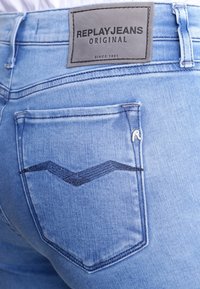 Jeans in denim azzurro chiaro con un design distintivo delle tasche, caratterizzati da cuciture a zigzag blu scuro e un'etichetta in pelle grigia sulla vita.