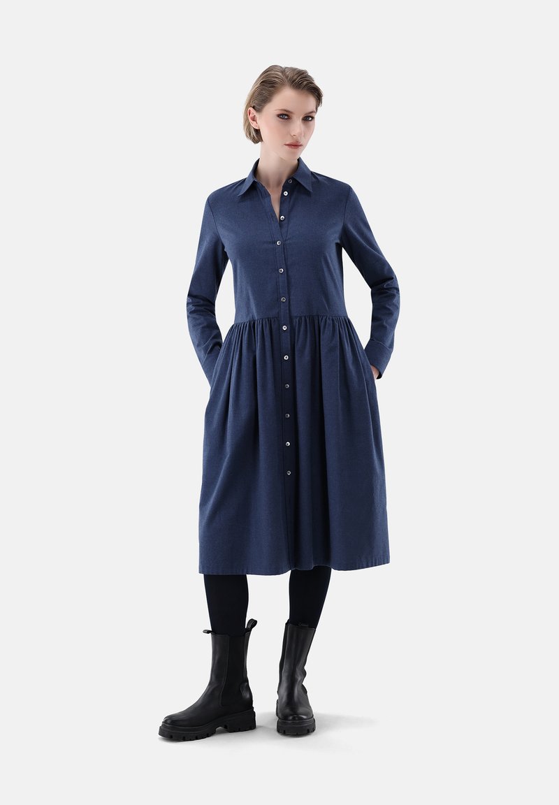 Knopfhemdkleid aus dunkelblauem Stoff, mit langen Ärmeln, tailliertem Schnitt und plissiertem Rock, sowie Seitentaschen, kombiniert mit schwarzen Ankle Boots.