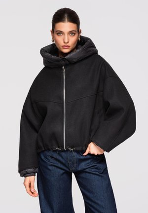 Femme portant une grande veste zippée noire avec un col rembourré et un jean bleu foncé, debout devant un fond gris clair.