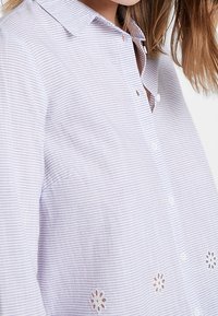 Hellblaues Button-Up-Hemd mit weißen Streifen und floralem Spitzenstickereien am Saum. Es hat einen Kragen und lange Ärmel.